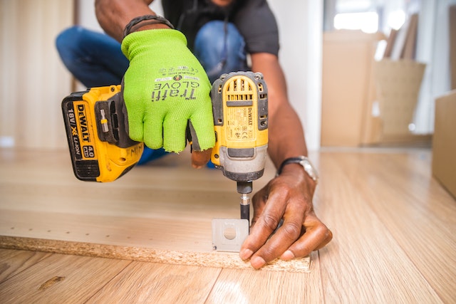 a carpenter in green work gloves uses a power drill to replace a bracket on a shelf in a rental property